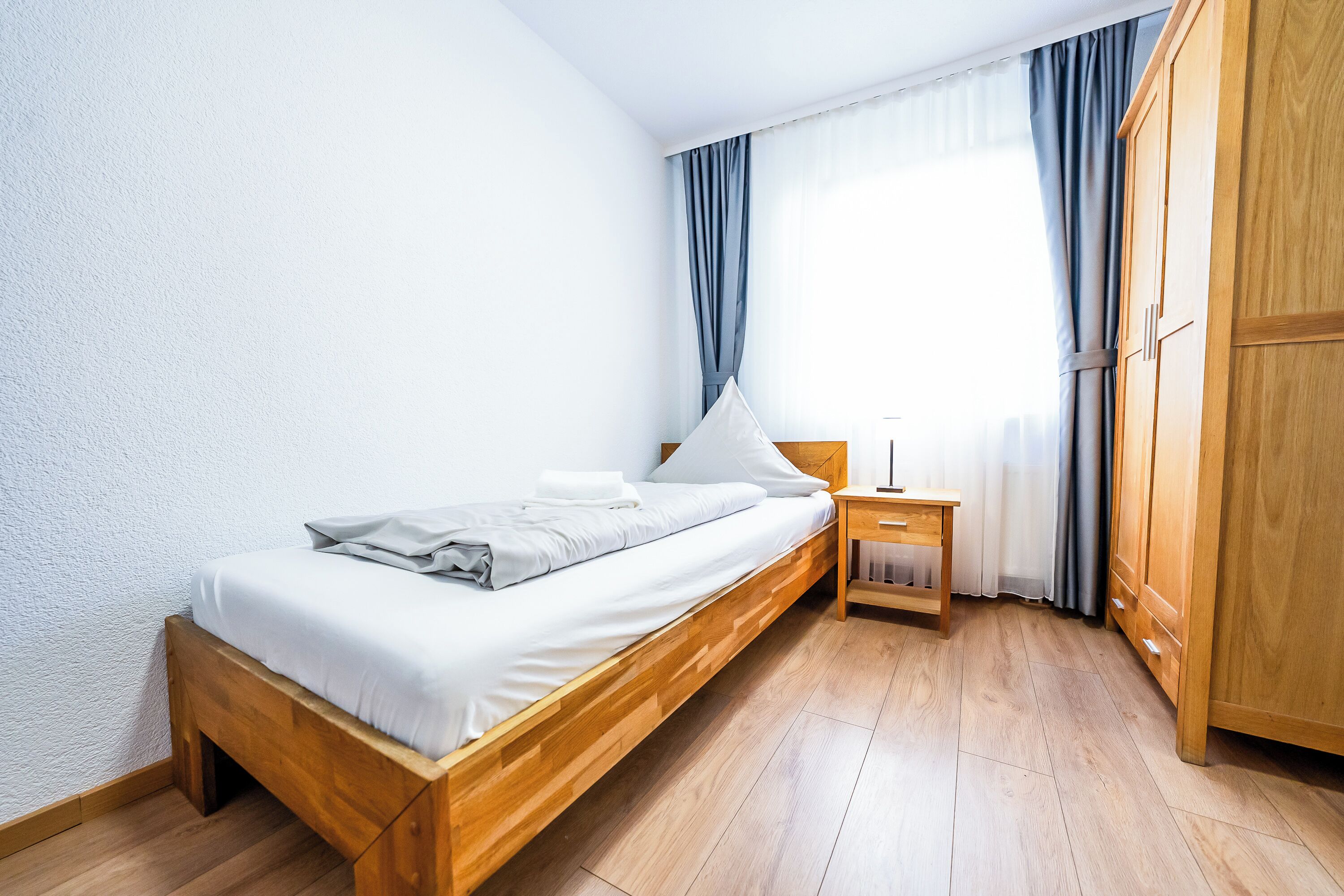 Una cama individual con estructura de madera, una mesita de noche y un armario en una habitación con suelo de madera y paredes blancas.