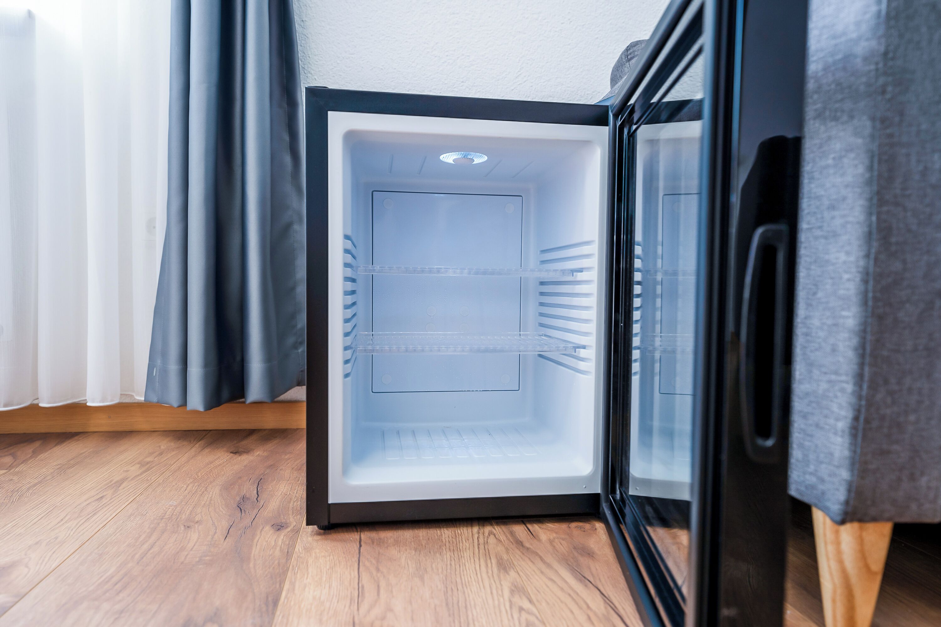 Un refrigerador compacto y moderno con puerta transparente, ubicado en un rincón de una habitación con piso de madera y pared blanca.