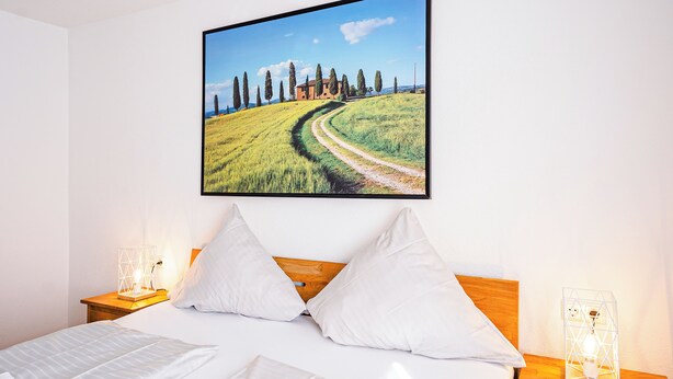 A bedroom with a bed, white pillows, a wooden headboard, a nightstand with a lamp, and a framed landscape painting on the wall.