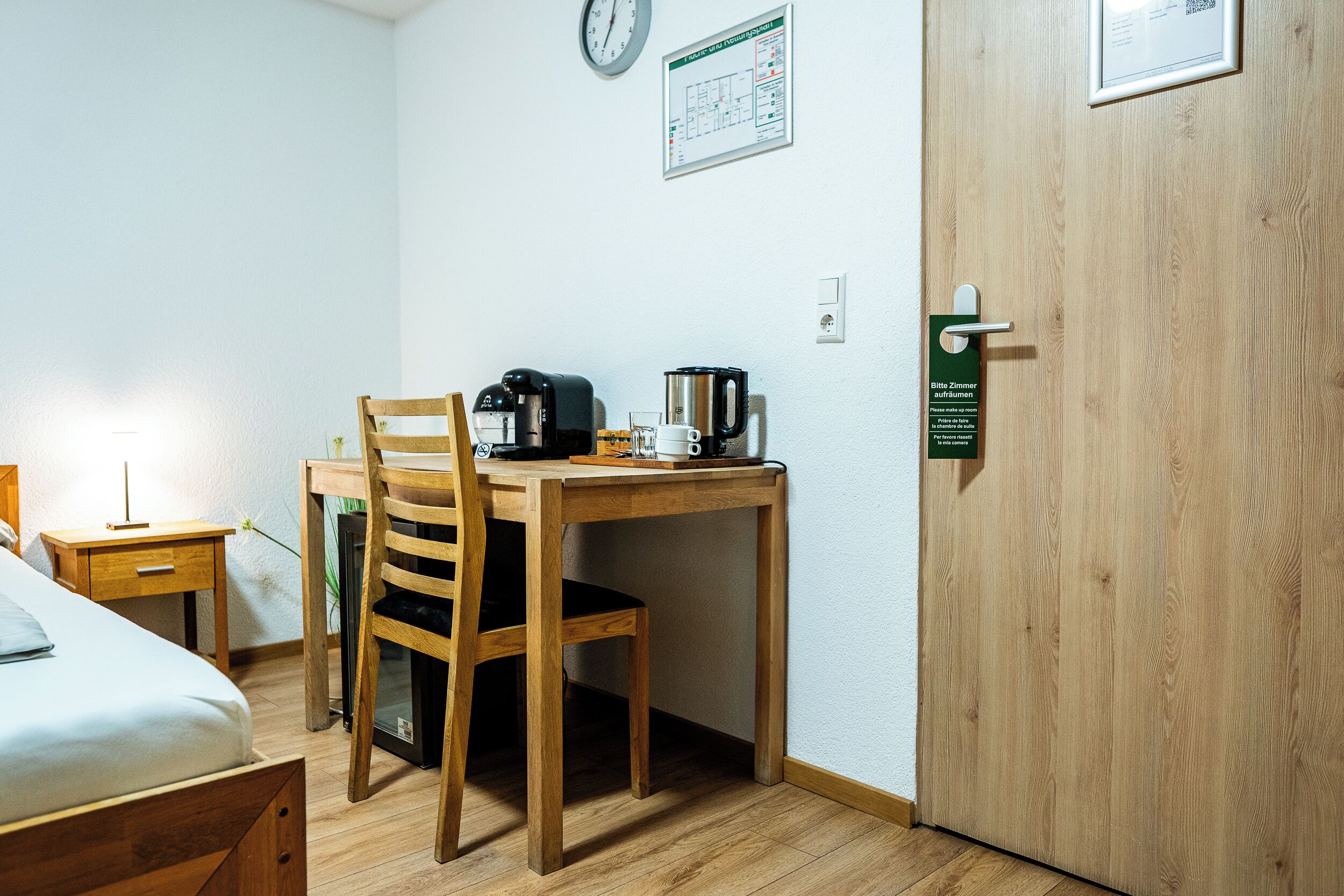A hotel room with a wooden desk, chair, and a small table with a kettle and coffee maker. There is a bed with a white comforter and a nightstand with a lamp.