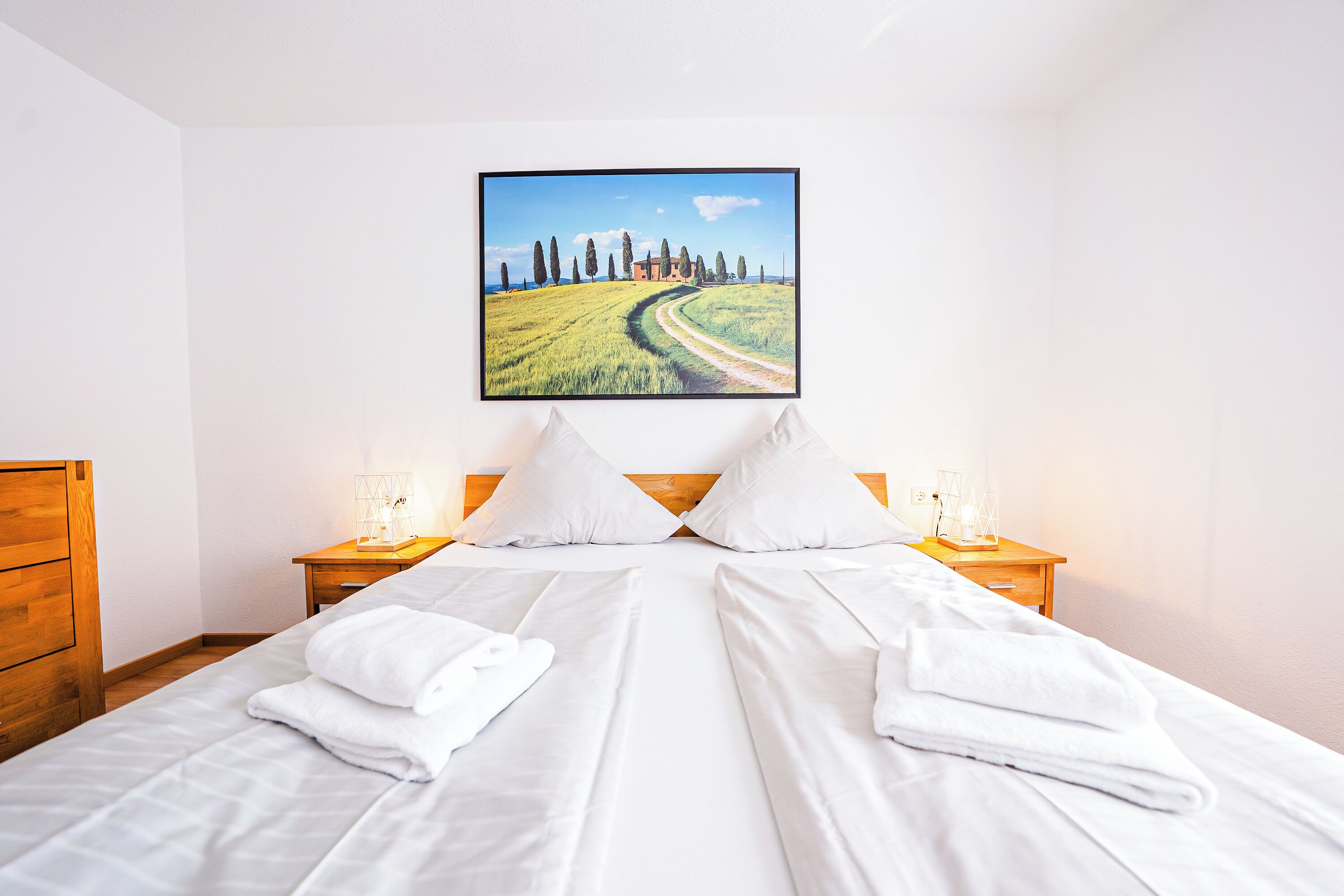 Habitación de hotel con una cama grande, dos mesitas de noche con lámparas, un aparador de madera y un cuadro con un paisaje en la pared.