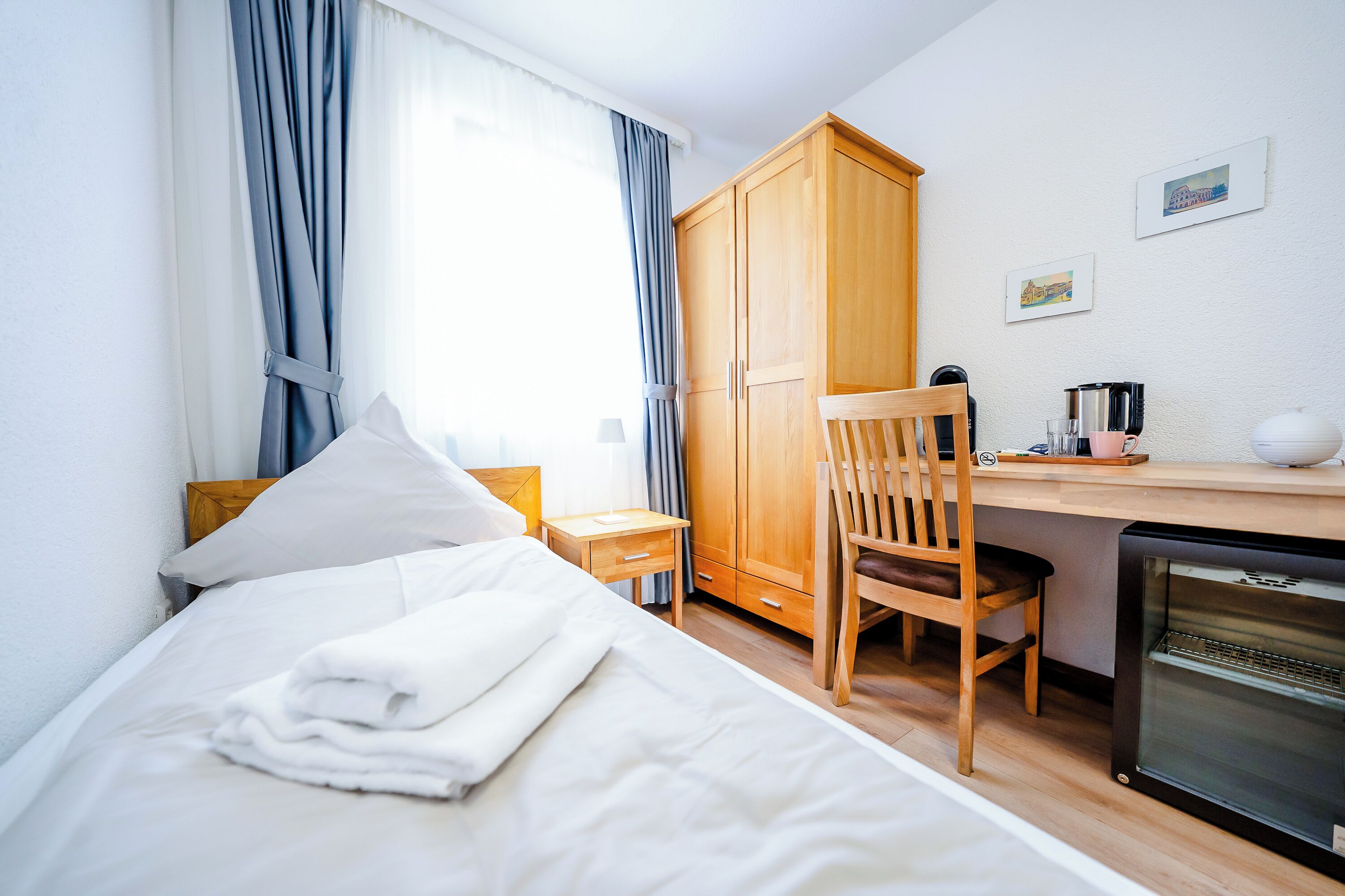 A bedroom with a bed, a wooden wardrobe, a desk with a chair, a coffee maker, and a refrigerator.