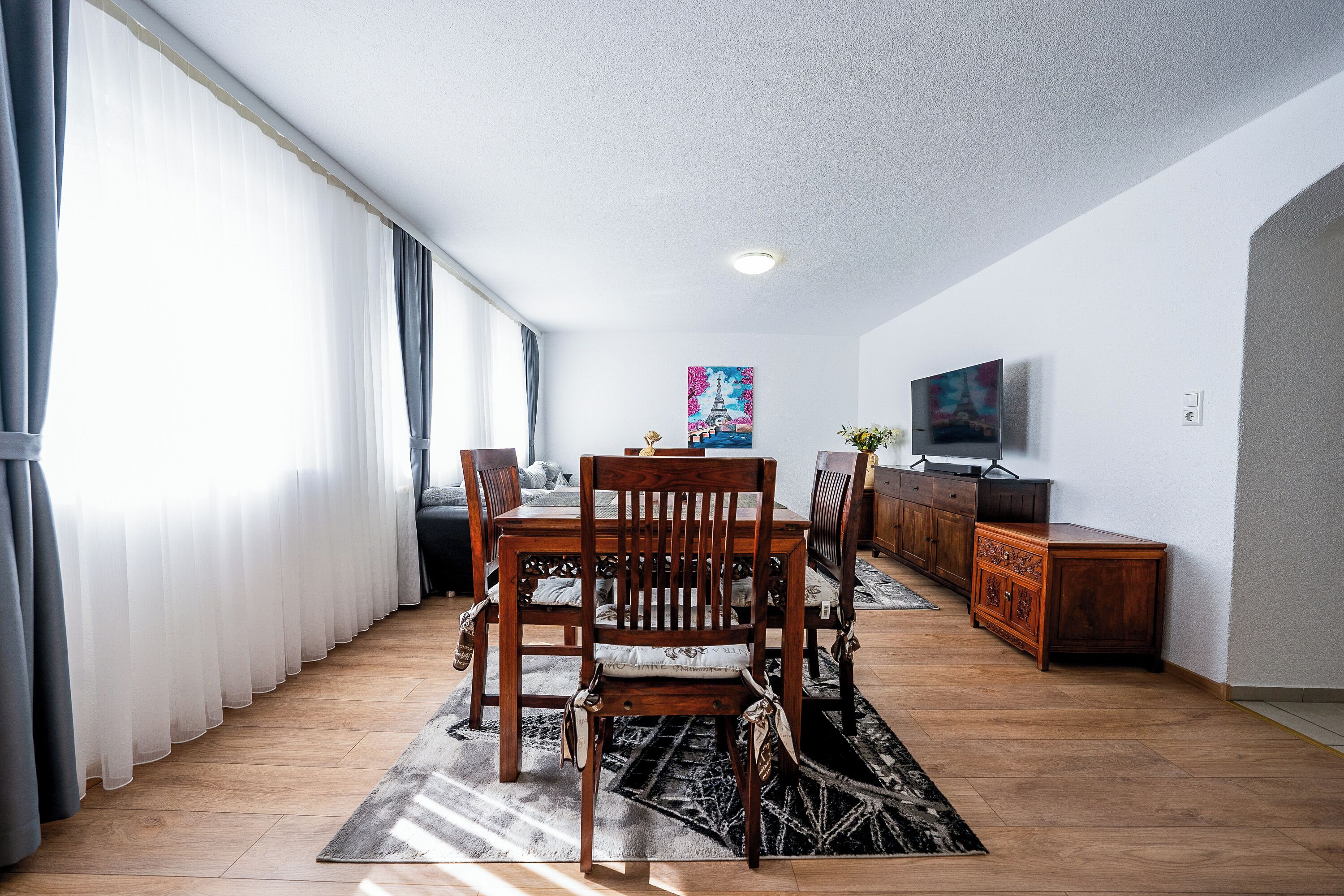 Un coin repas avec une table en bois, des chaises et un tapis. Il y a un téléviseur accroché au mur et un tableau au-dessus.