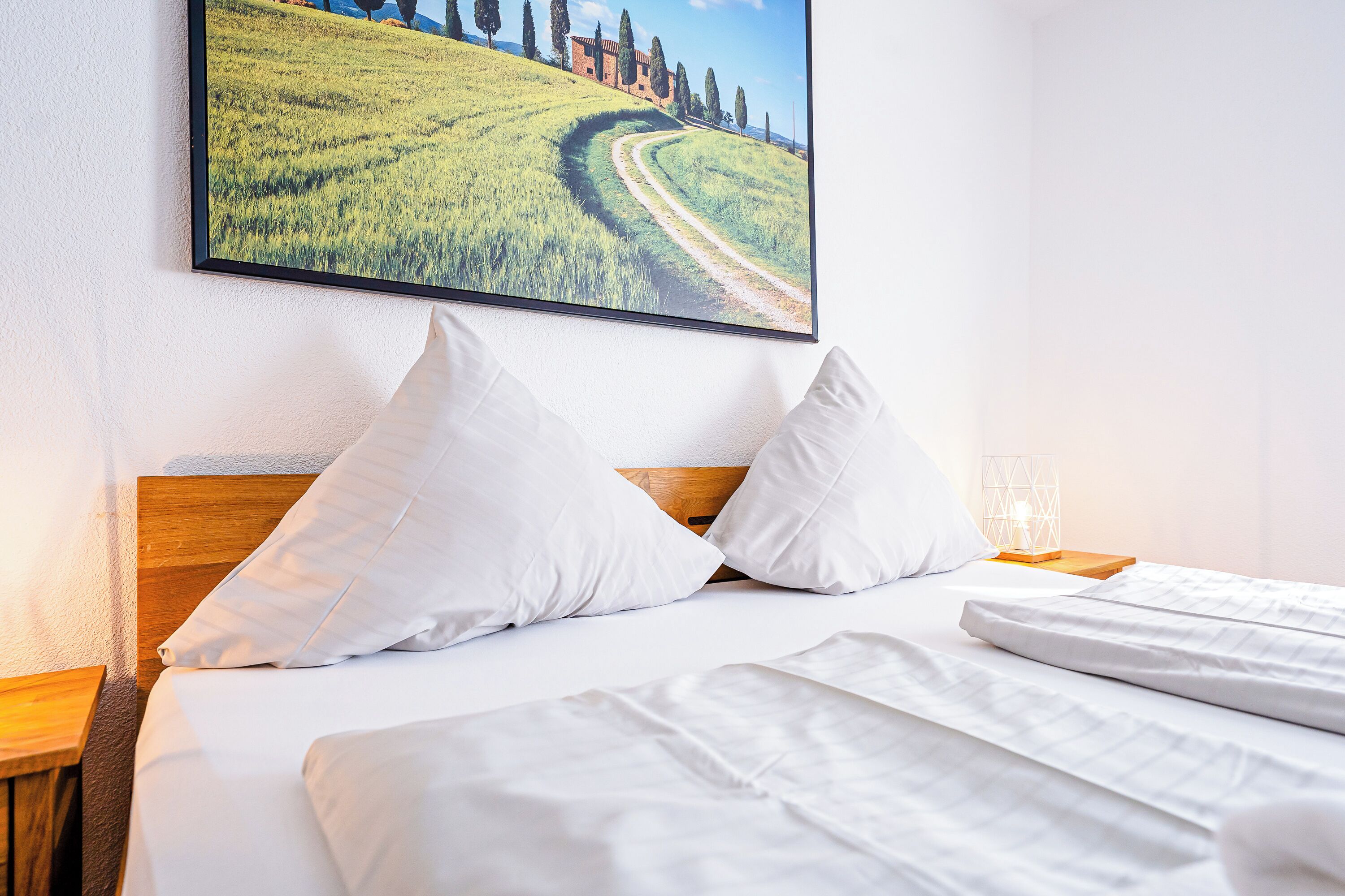 A neatly made bed with white linens and pillows, a wooden headboard, and a framed picture of a countryside landscape on the wall.