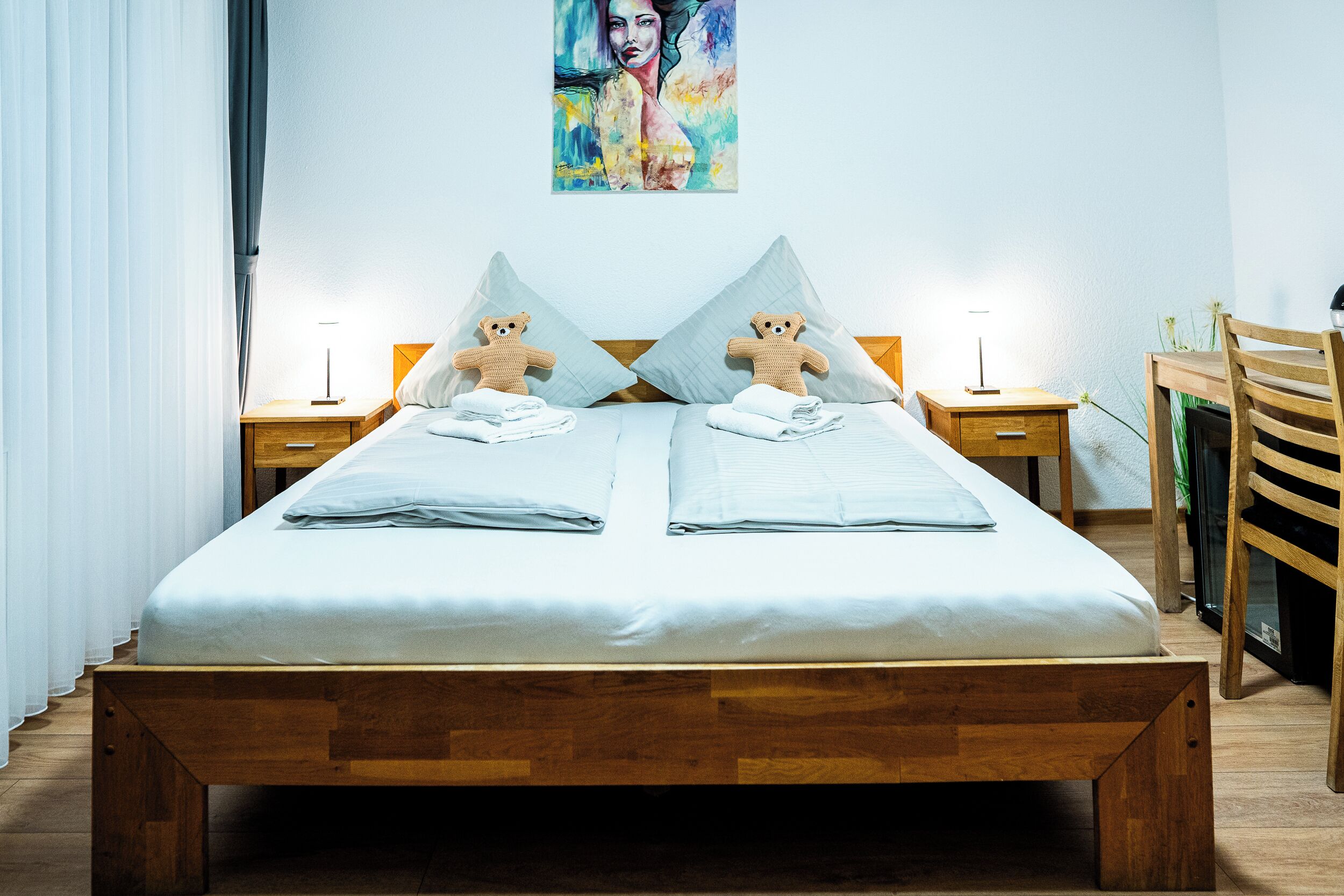 Una cama doble con dos almohadas, cabecera de madera, mesita de noche con lámpara y una silla.