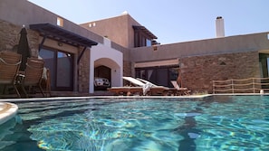Une piscine avec des chaises longues et des parasols, un mur en pierre et un véhicule garé à proximité.