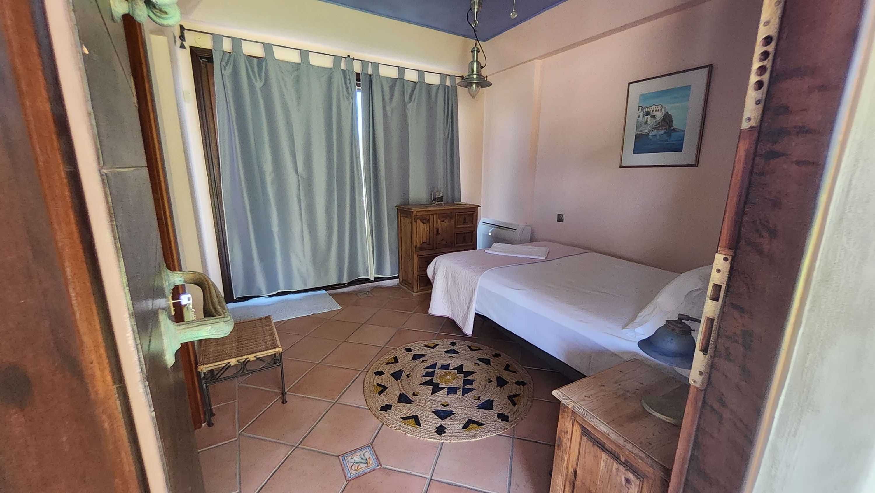 A bedroom with a bed, a wooden nightstand, a small table, a chair, a window with curtains, and a painting on the wall.