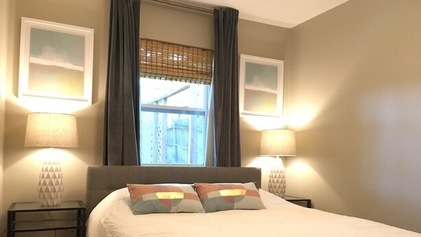 A bedroom with a large bed, two bedside lamps, a window with curtains, and two wall-mounted picture frames. - Charming 4BR Duplex Home |  5&6 (Kansas City)