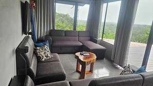 A modern living room with a sectional sofa, a small coffee table, and large windows offering a view of greenery. - Welcome to Osa Views we’re really happy to have you in our house . (Provincia de Puntarenas)