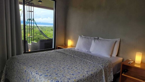 A bedroom with a large bed, a nightstand with a lamp, and a view of a lush landscape through a sliding door. - Welcome to Osa Views we’re really happy to have you in our house . (Provincia de Puntarenas)