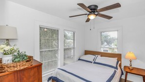 Ein Schlafzimmer mit Bett, Holz-Kopfteil, zwei Fenstern mit Jalousien, Deckenventilator, Nachttisch mit Lampe und einer Holzkommode mit einem Blumenkorb.