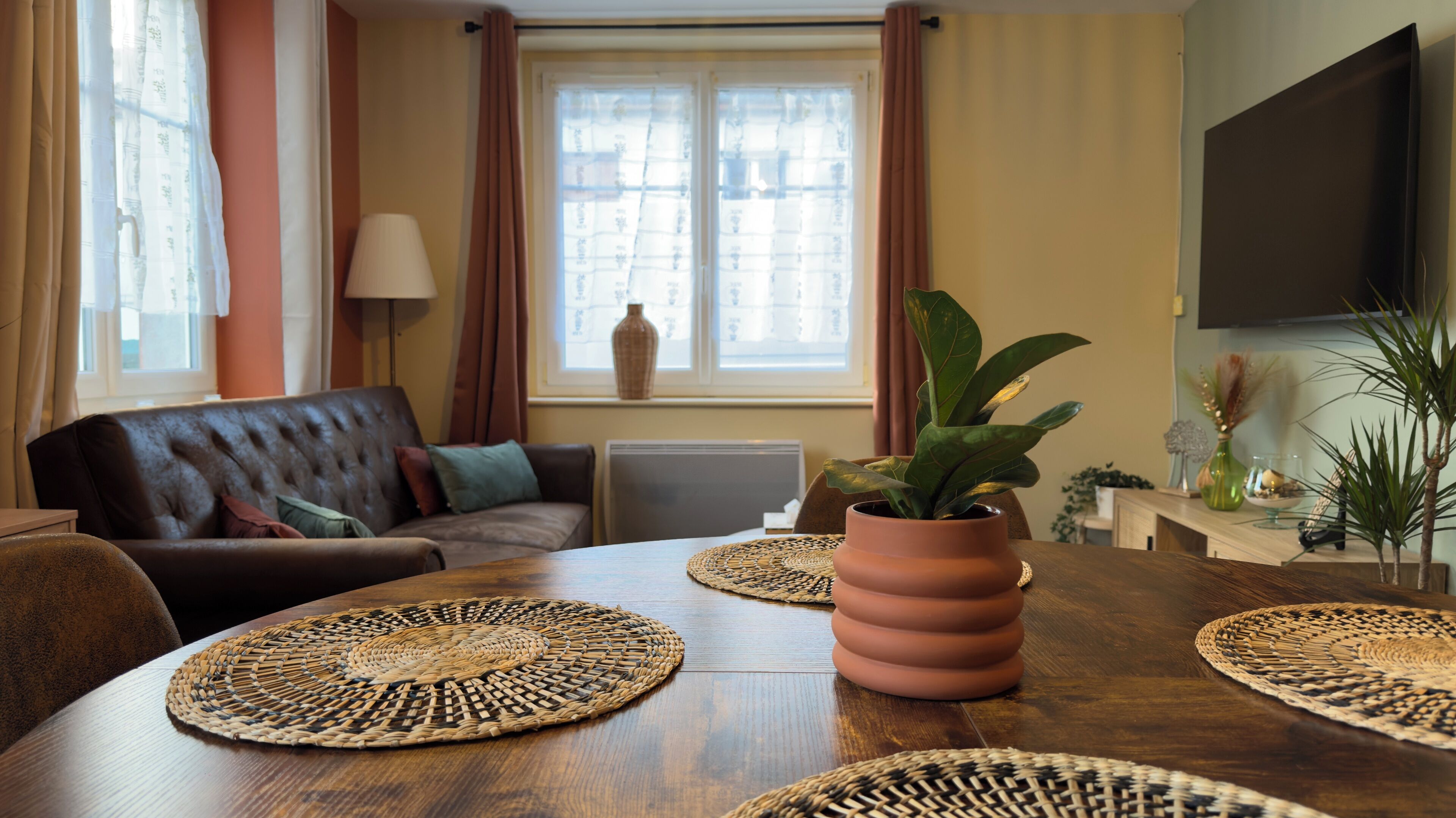 A living room with a brown leather sofa, a wooden dining table with woven placemats, a potted plant, and a large flat-screen TV mounted on the wall.