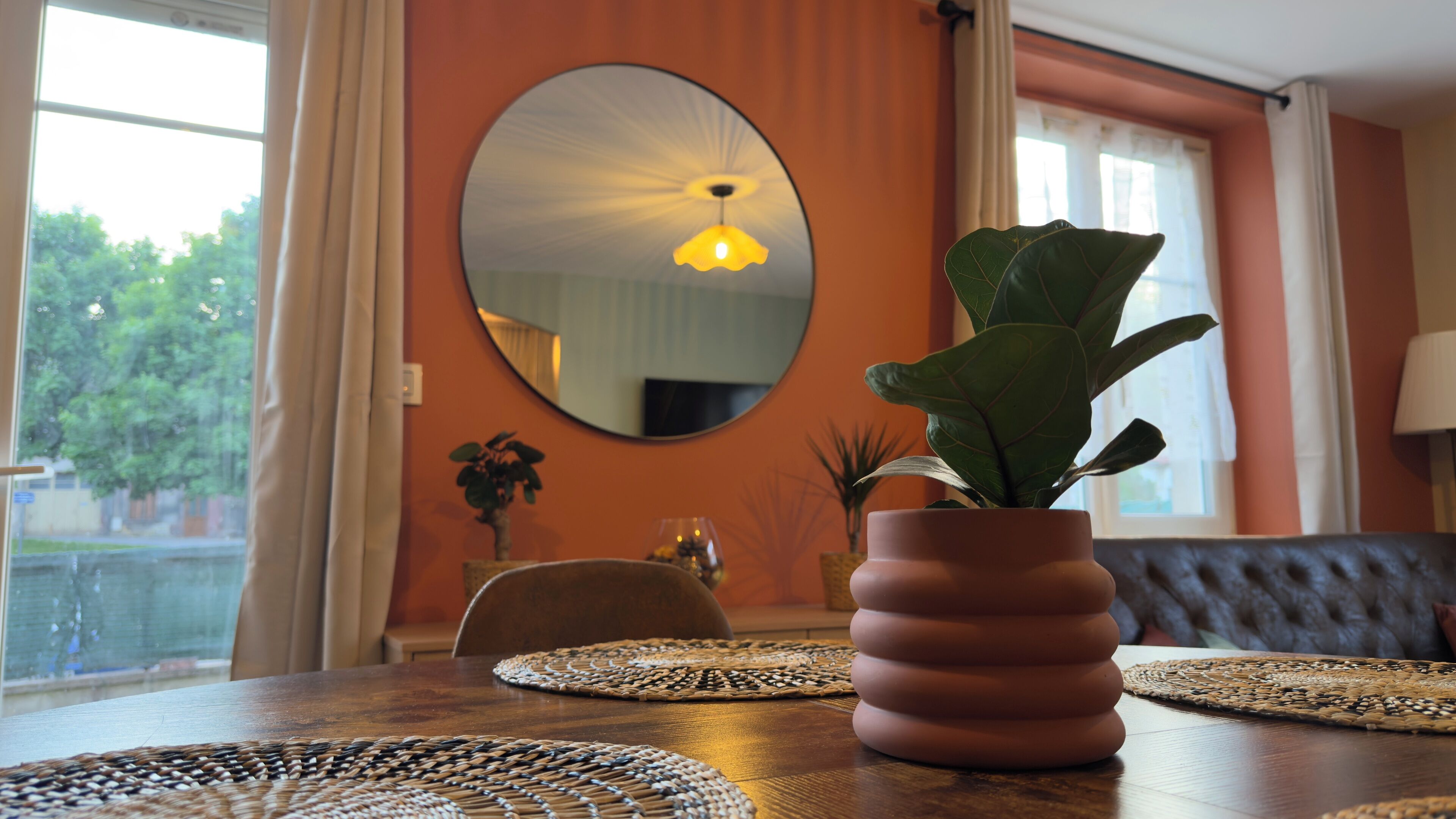 Een eethoek met een ronde spiegel, een potplant en een tafel met geweven placemats.