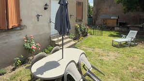 En terrasse med et rundt bord og stole, en lille parasol, samt en have med blomster og en liggestol.