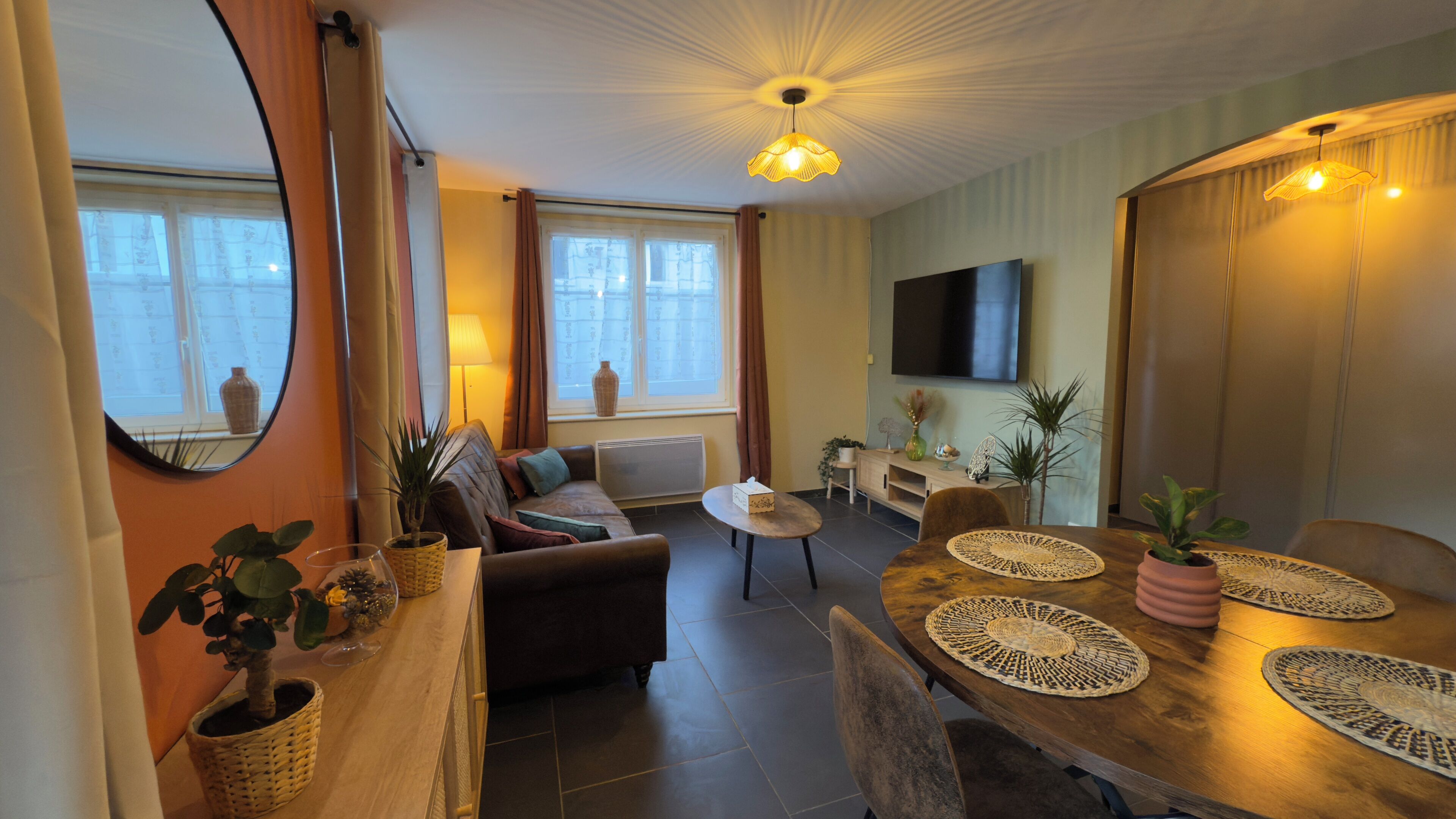 Un salon moderne comprenant un canapé, une table basse, une table à manger avec des sets de table, un téléviseur et une grande fenêtre avec des rideaux.