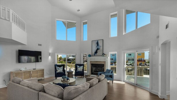 A spacious living room with a large sectional sofa, a flat-screen TV, and a fireplace. There are multiple windows providing natural light and views of the outside.