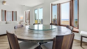 A spacious living room with a round glass-top table, wooden chairs, a white sofa, and large windows offering a view of the beach.