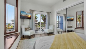 A hotel room with a large bed, two white armchairs, a TV, and a balcony with a view of palm trees and buildings.