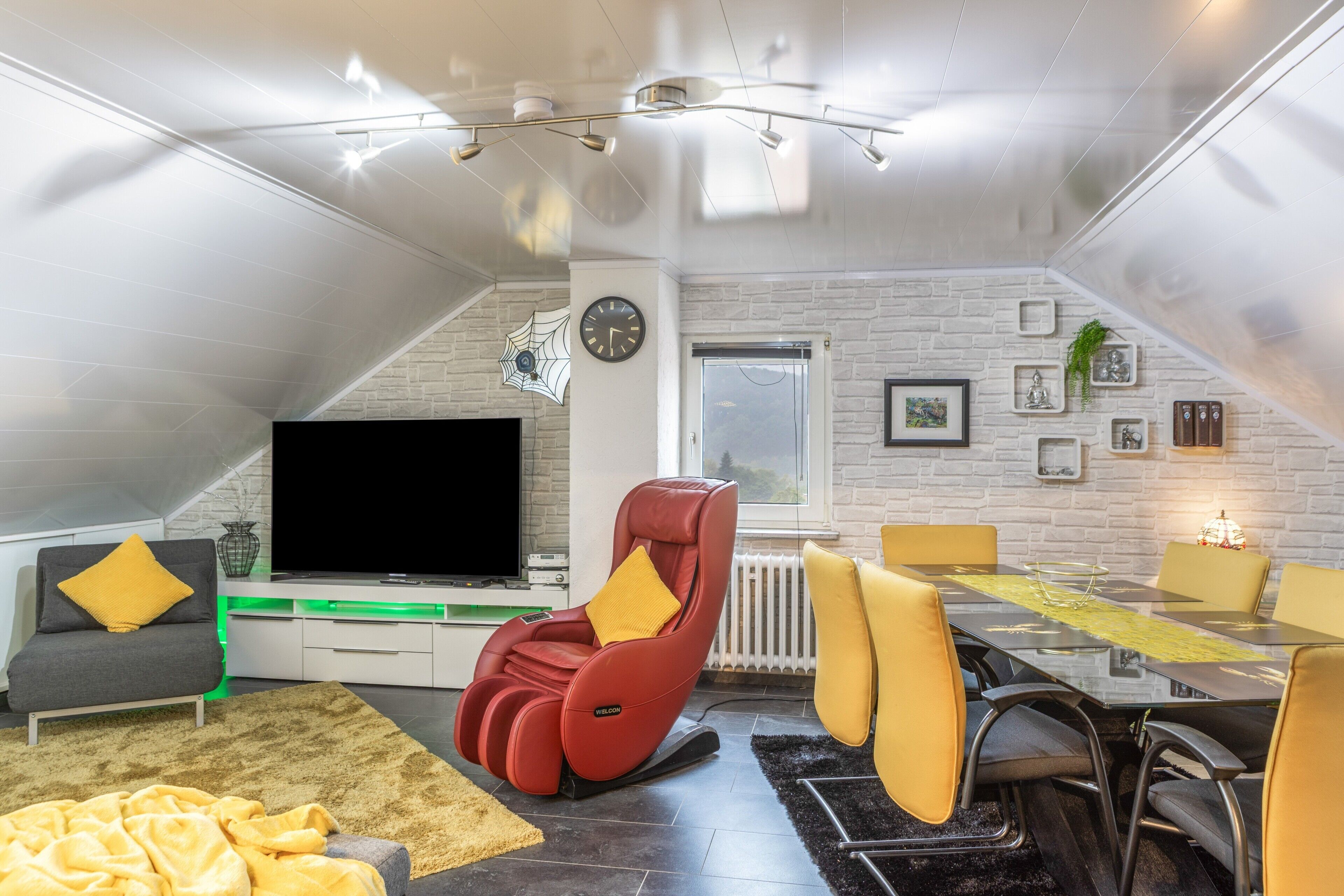 Un salón moderno con televisor de pantalla plana, una silla de masaje y un comedor con sillas amarillas.
