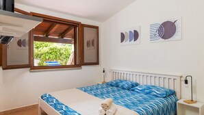 Ein Schlafzimmer mit Bett, Fenster, einem an der Wand befestigten Fernseher und einem Nachttisch mit Lampe.