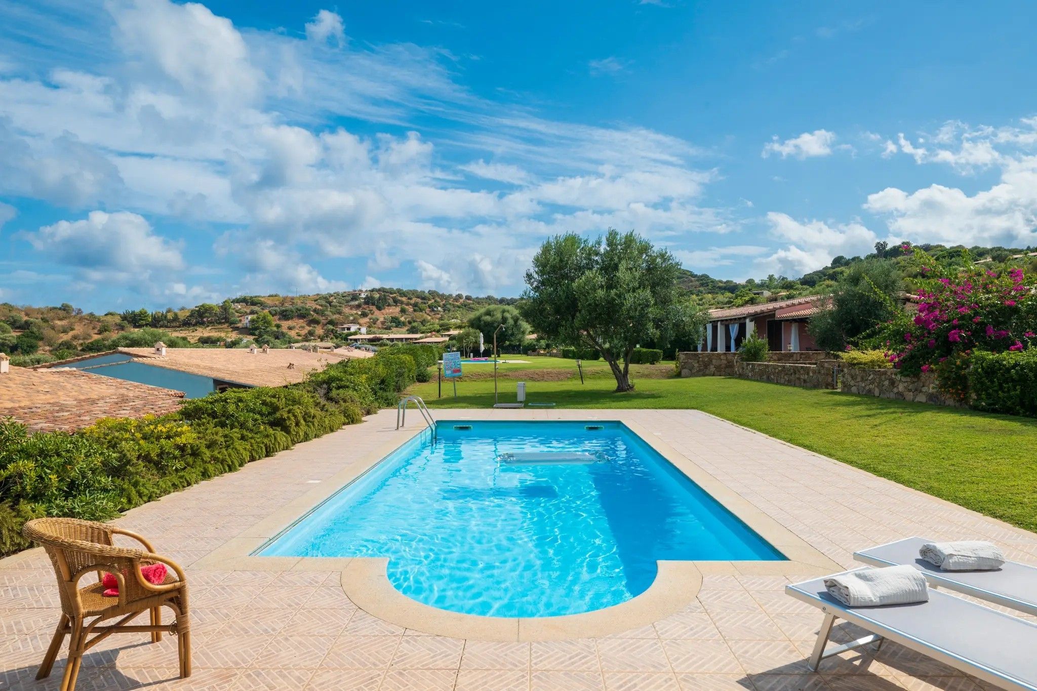 Une piscine rectangulaire à l’eau d’un bleu limpide, entourée d’une terrasse dallée. On y trouve des chaises longues et un fauteuil en osier avec un coussin. La piscine est mise en valeur par un ciel dégagé et un paysage vallonné verdoyant en arrière-plan.