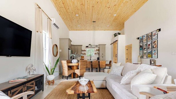 A modern living room with a wooden ceiling, a large flat-screen TV, a dining area with wicker furniture, and a sofa with decorative pillows. - Rustic Elegance Farmhouse for a tranquil retreat experience in Sanford, FL (Sanford)