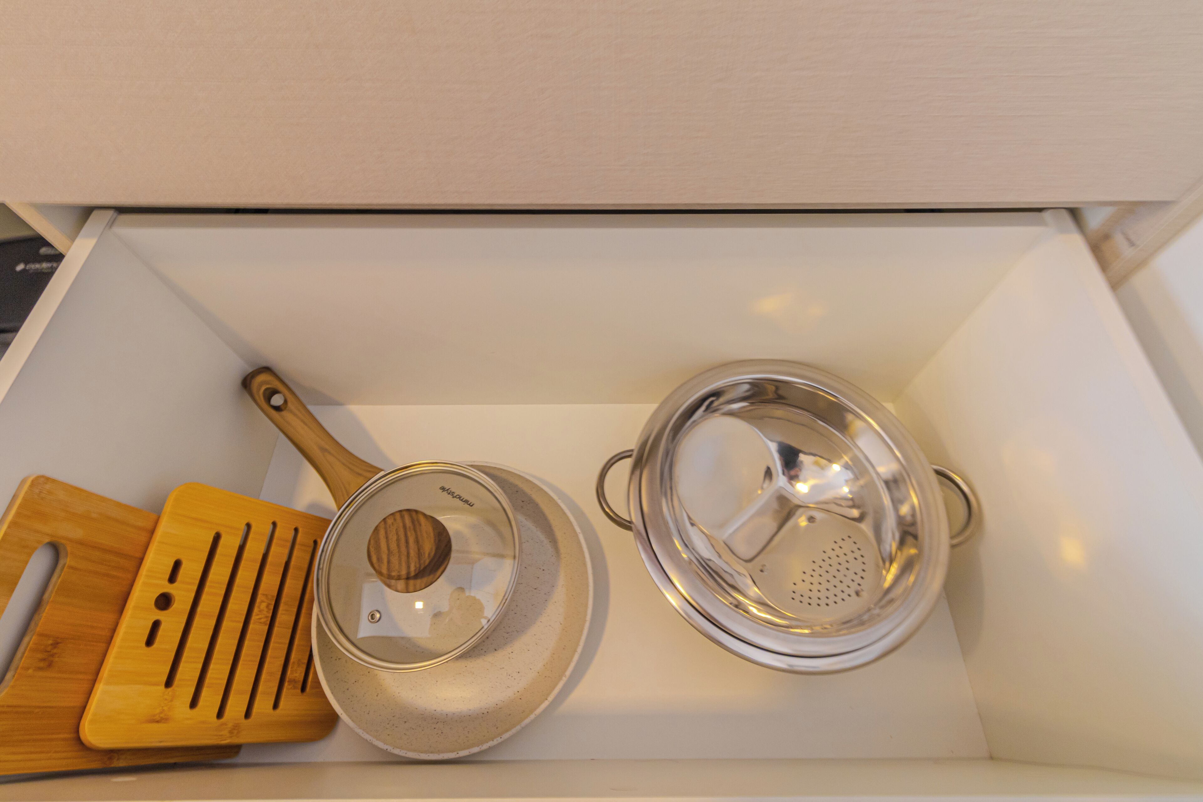 A kitchen drawer containing a wooden cutting board, a metal pot with a lid, and a metal colander.