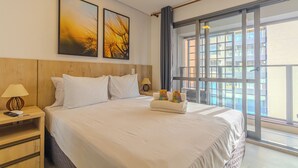 Une chambre d’hôtel avec un grand lit, deux œuvres d’art accrochées au mur, une table de chevet avec une lampe et une vue sur la ville à travers de grandes fenêtres.