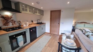 A compact kitchen with a gas stove, oven, and a small refrigerator. There is a dining area with a table set for two, and a sofa in the adjacent living room. - Bailem Lodge at Loch Ness (adults only) (Fort Augustus)