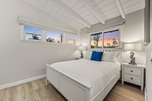 A bedroom with a large bed, two bedside tables, and a window with a view of palm trees and a sunset.