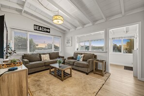 A spacious living room with two sofas, a coffee table, and a TV. There are large windows with blinds, a ceiling fan, and a sign that reads "BEACH". - Perfect Location, Amazing Reviews, Walk To Beach (Ventura)