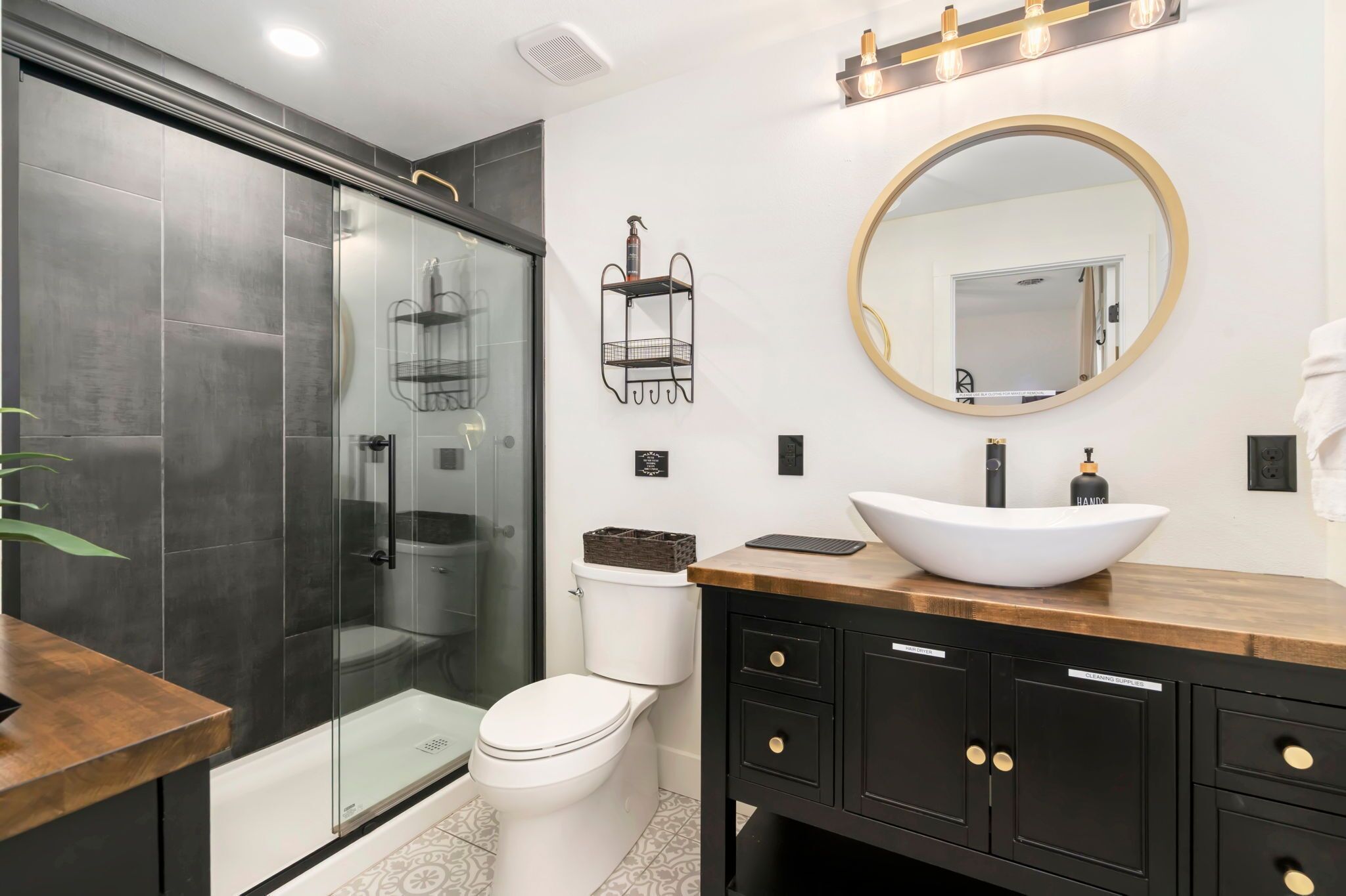 A modern bathroom with a glass-enclosed shower, a white toilet, a wooden vanity with a large mirror, and a black cabinet with gold handles.