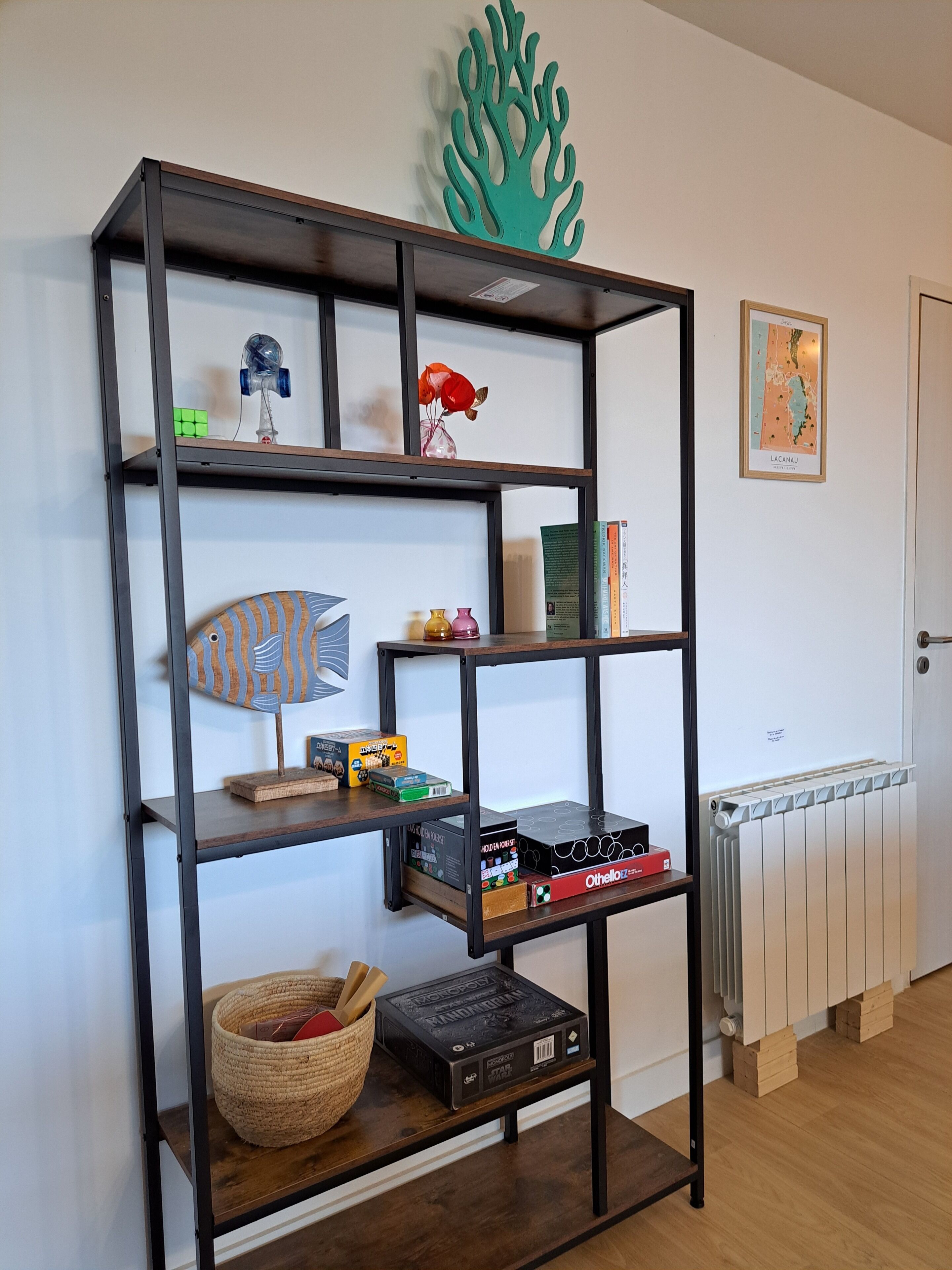 Une pièce avec une étagère en métal noir adossée à un mur blanc, contenant divers objets tels que des jeux de société, une petite plante et des objets décoratifs.
