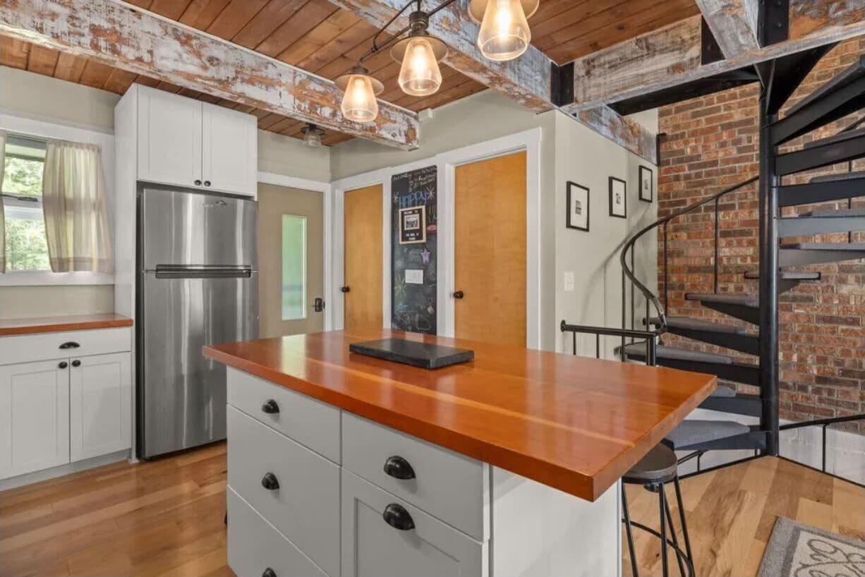 A kitchen with white cabinets, a stainless steel refrigerator, a wooden countertop, and a black island. There is a staircase with a metal railing leading to a brick wall.