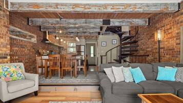 A spacious living area with a gray sofa, a wooden coffee table, and a colorful chair. A staircase leads to an upper level, and a dining area is visible in the background.