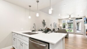A modern kitchen with white cabinetry, stainless steel appliances, pendant lighting, and a dining area with a sofa and table. - Northfield Old Town Flat Attached Garage w/ EV (Fort Collins)