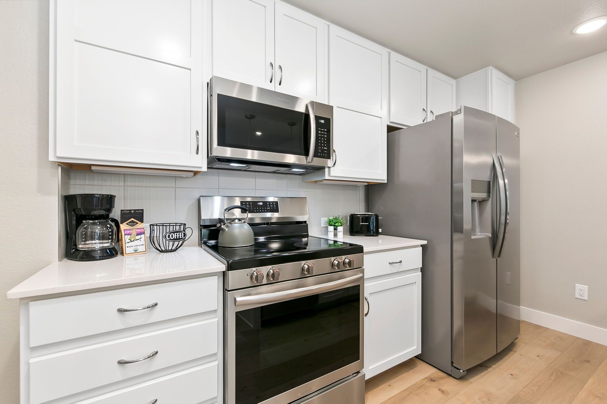 A modern kitchen with white cabinets, a stainless steel refrigerator, a built-in oven, a microwave, and a coffee maker.
