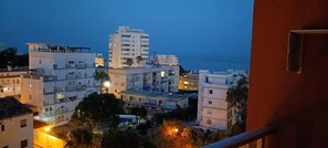 Un paisaje urbano costero al atardecer, con edificios de varios pisos, una calle visible y una masa de agua en el fondo.