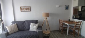 Un salón con un sofá gris, una mesita redonda pequeña, una lámpara y una zona de cocina con armarios y un microondas.