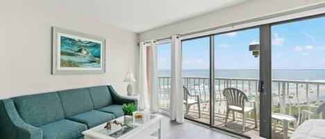 Een moderne woonkamer met een blauwgroene bank, een witte salontafel en een grote glazen deur die uitkomt op een balkon met uitzicht op het strand.
