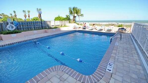 Une piscine rectangulaire dotée de couloirs délimités, d’un poste de surveillance pour les sauveteurs et d’une zone de détente avec des chaises longues, offrant une vue sur la plage.