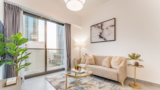 A modern living room with a sofa, a glass coffee table, a floor lamp, a potted plant, and a view of a cityscape through large windows. - Near Circle Mall And Parks, Easy Access to Al Khail Road (Dubai)