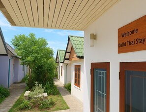 Un pasillo con una puerta de madera a la derecha, un letrero que dice “Welcome Dalbt Thai Stay” y un sendero que conduce a un edificio con techo verde.