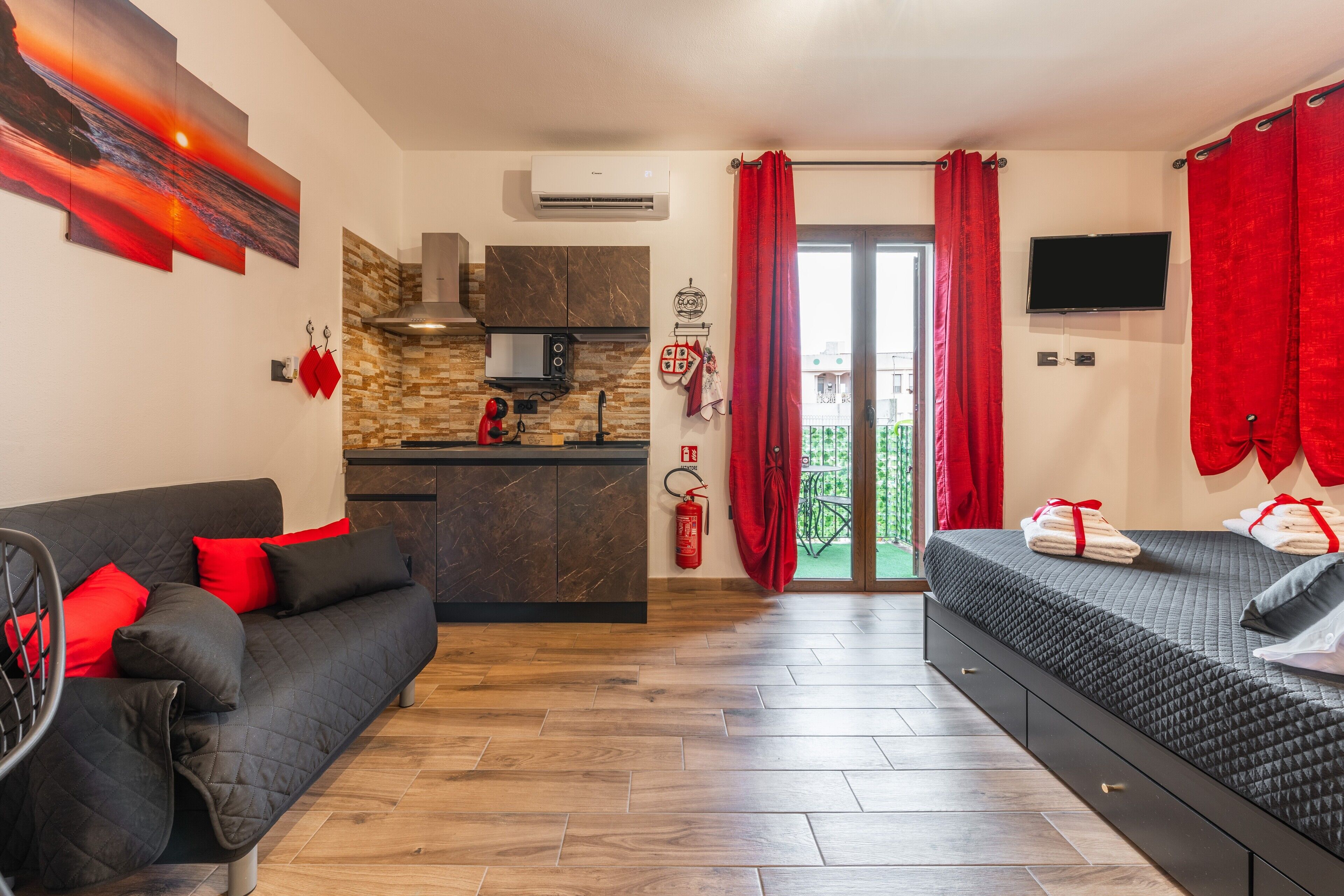Una habitación moderna con sofá, cama, zona de cocina y balcón con cortinas rojas.