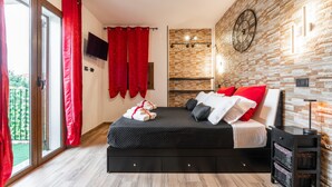 A modern bedroom with a bed, red curtains, a TV, a brick wall, and a wooden floor. - Studio 'Attilio's House' with Balcony, Wi-Fi and Air Conditioning (Capoterra)