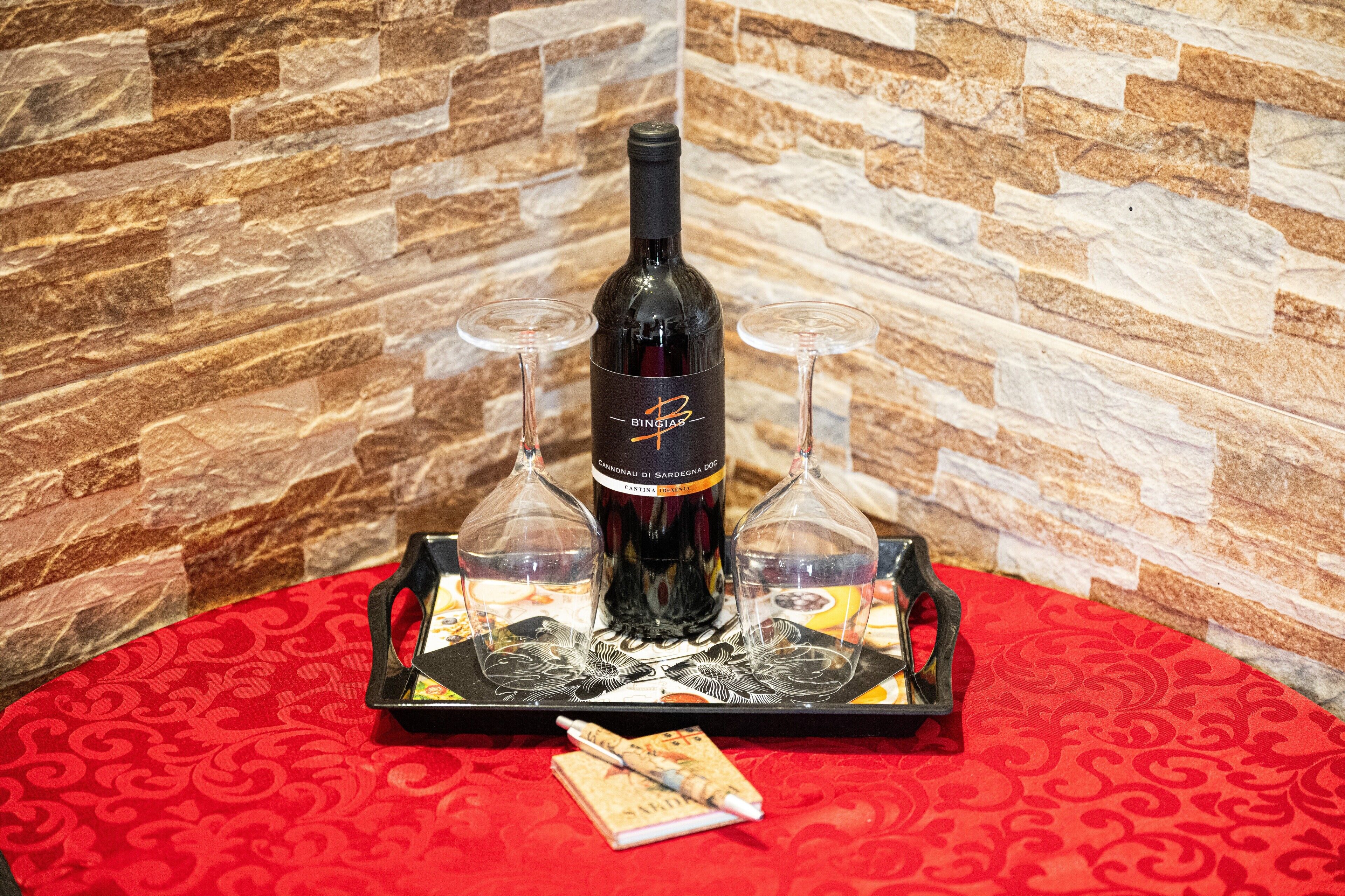 Una bottiglia di vino su un vassoio con due bicchieri, un taccuino e una penna, il tutto su una tovaglia rossa.