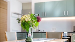 Une table à manger ornée d’un vase de fleurs, des chaises en bois et un coin cuisine avec des placards en arrière-plan.