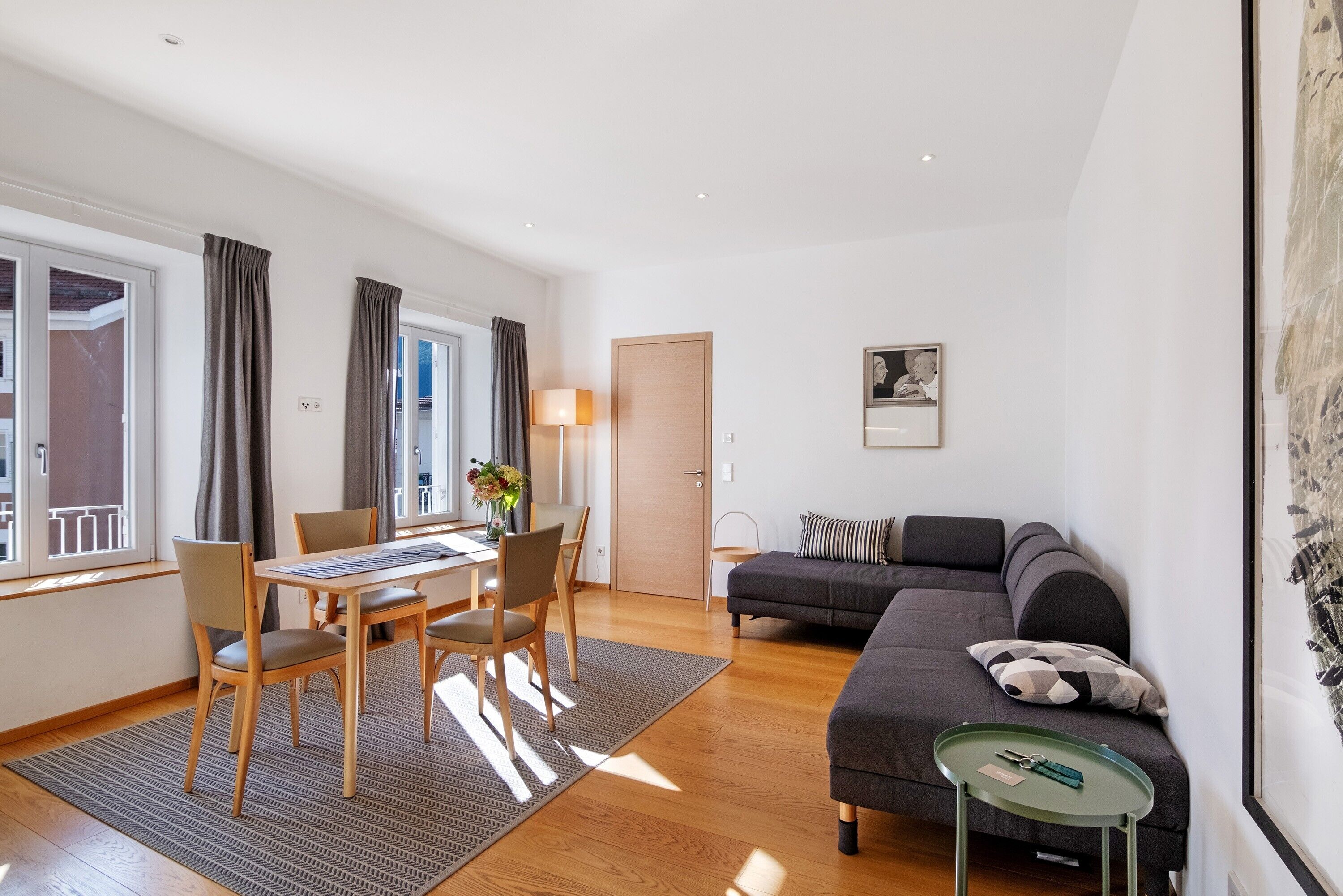 A modern living room with a sofa, a dining table with chairs, a small side table, and a door leading to another room.