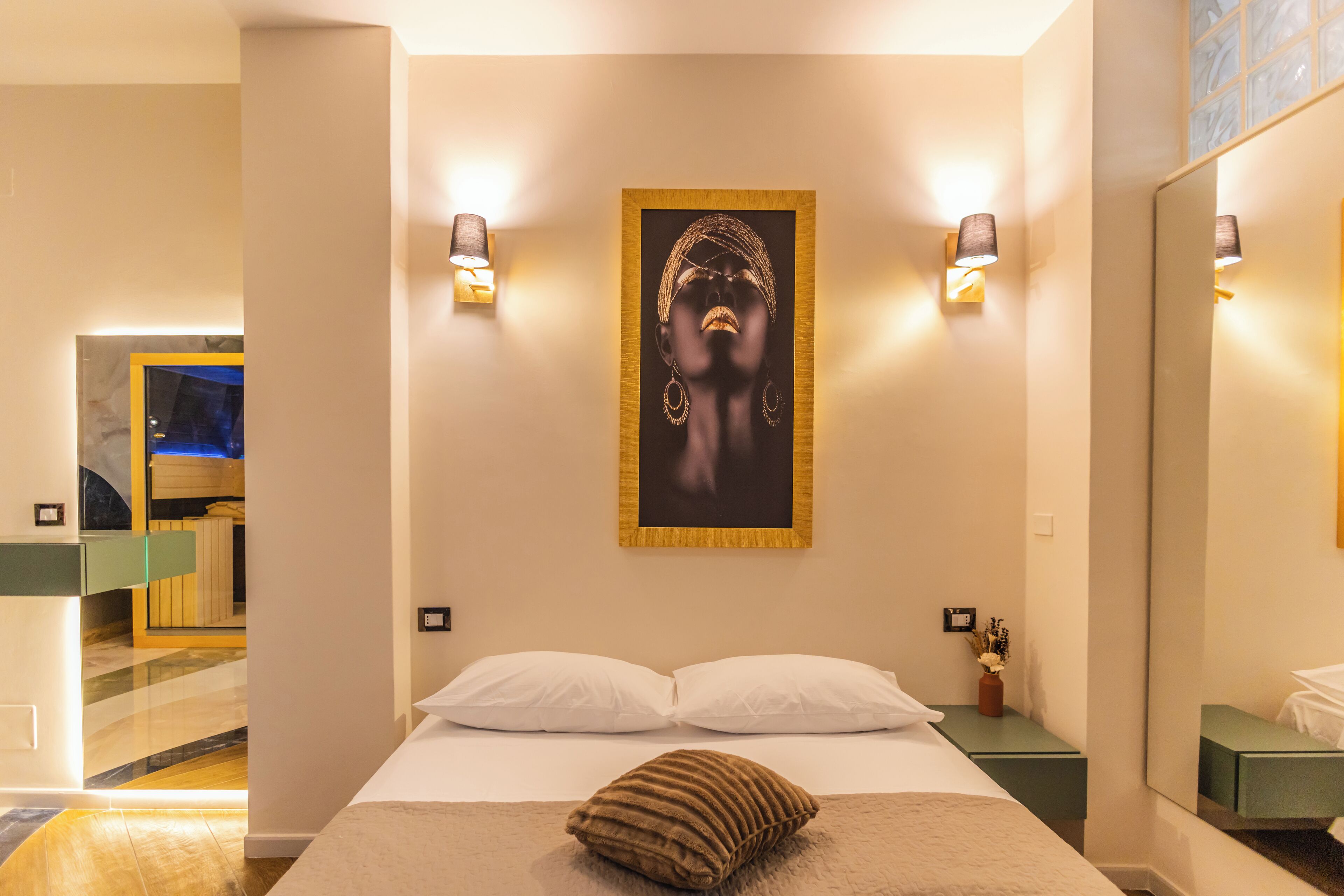 A hotel room with a bed, pillows, a framed artwork, bedside lamps, a mirror, and a small table with a vase.