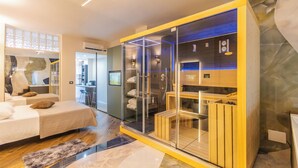 Habitación de hotel moderna con zona de ducha de vidrio, un banco de madera y un rincón de descanso con vistas a la cocina.